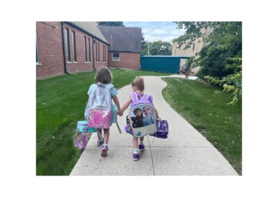 Colorful preschool backpacks on two young girls walking on a curved sidewalk at Montessori Stepping Stones daycare in Mount Clemens, MI, demonstrating early childhood education and nurturing environment.