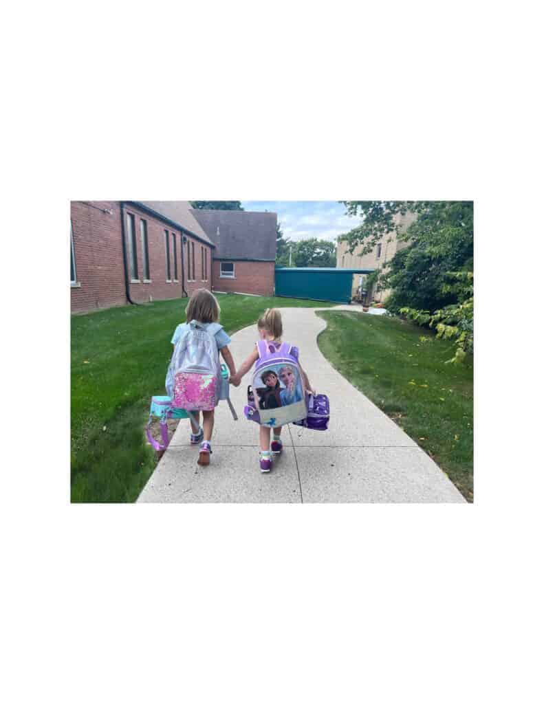 Brightly colored backpacks on two young girls walking hand in hand toward Montessori preschool in Mount Clemens, Michigan, illustrating a caring and nurturing daycare environment for early childhood education.