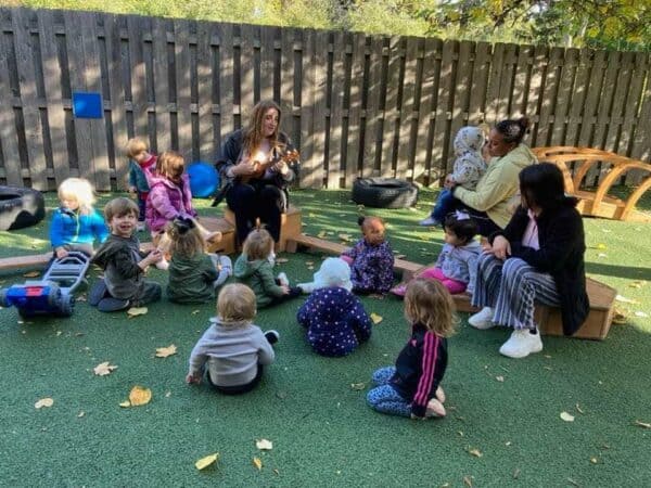 Teacher playing the ukulele for children in the Montessori toddler program during an outdoor music circle time session.