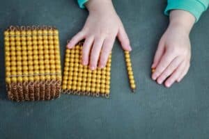 Top-down view of a child's hands working with Montessori golden beads, showing a thousand cube, a hundred square, a ten bar, and a single unit bead on a green work mat to learn the decimal system and place value.