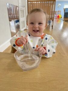 Cheerful toddler happily stirs batter in a clear glass bowl at Montessori Stepping Stones, demonstrating early sensory and motor skills development in a nurturing learning environment.
