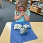 Preschool student using a magnifying glass to examine fossils and rocks during a Montessori science nature lesson.