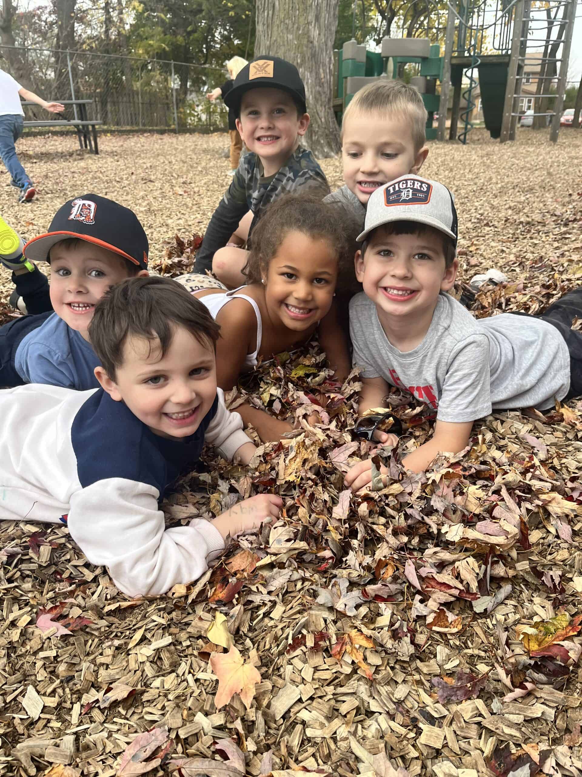 A diverse group of smiling Montessori preschool children lying together in a large pile of autumn leaves on the school playground, demonstrating social connection and outdoor exploration.