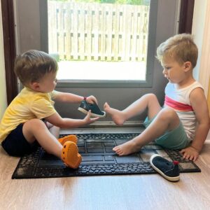 Two young Montessori students sitting on a mat helping each other put on shoes, demonstrating social skills and independence at Montessori Stepping Stones in Macomb County.