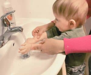 A toddler standing at a sink receiving gentle guidance from a teacher on how to wash hands as part of the Montessori practical life curriculum.