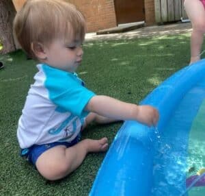 Toddler sitting on the grass splashing in a blue water pool during Montessori summer program sensory play.